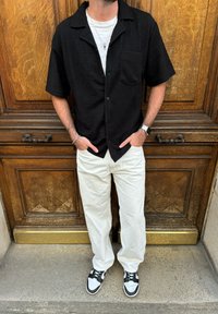 Μαύρο κοντομάνικο πουκάμισο με υφή και τσέπη στο στήθος, συνδυασμένο με λευκά φαρδιά παντελόνια και μαύρα-άσπρα αθλητικά παπούτσια.