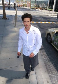 Camisa branca de botões com detalhes bordados, combinada com calças largas cinzas e sapatos pretos robustos. Ambiente ao ar livre com calçada e edifícios.