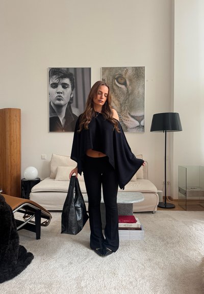 Mujer vestida de negro, sosteniendo una gran bolsa negra, de pie en una sala de estar con un sofá beige, dos fotos enmarcadas, una lámpara de pie y una silla moderna.