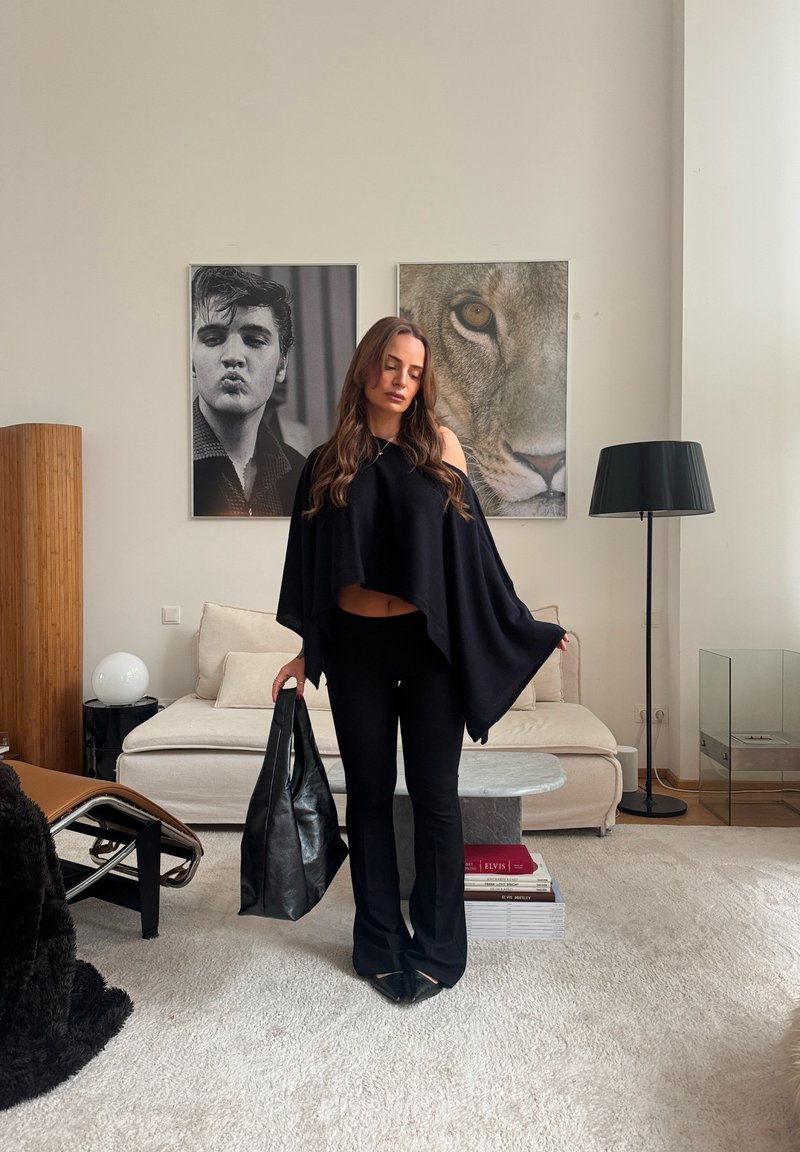 Fekete ruhát viselő nő, kezében egy nagy fekete táskával, egy nappaliban áll, ahol bézs kanapé, két keretezett fénykép, állólámpa és modern szék található.