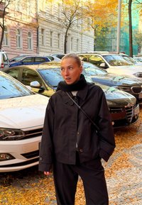 Černá bunda s velkými knoflíky a volným střihem, doplněná o černé kalhoty ve stejném stylu. V pozad�í jsou auta a podzimní listí.