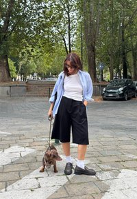 Culottes pretos, t-shirt branca e uma camisa azul às riscas. Sapatos castanhos e meias brancas. Um dachshund castanho com uma trela. Ambiente urbano ao ar livre.
