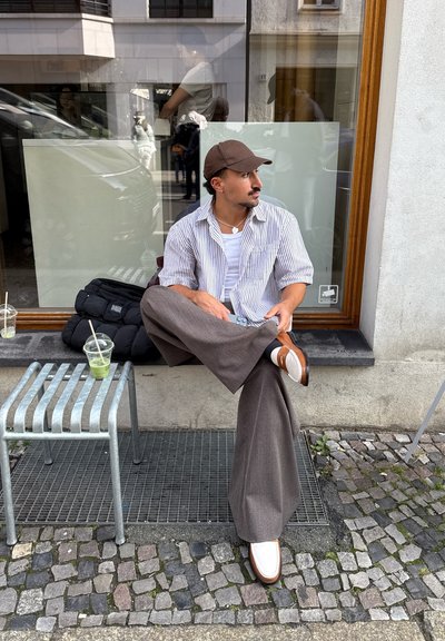 Persona sentada sobre una rejilla de metal, vistiendo una camiseta blanca a rayas sobre una camiseta de tirantes blanca, pantalones anchos grises y zapatos de slip-on marrones; dos bebidas cercanas.