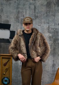 Veste courte en fausse fourrure marron avec capuche, portée sur une chemise noire et un pantalon taille haute marron ; une casquette marron ajoute une touche décontractée.