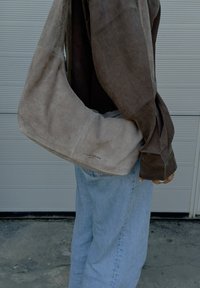 Bolso de hombro de ante beige con un diseño curvado, que presenta un logo de la marca visible en la parte inferior. Llevado con una camiseta de manga larga oscura y jeans claros.