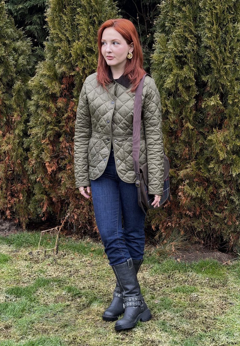 Femme aux cheveux roux portant une veste matelassée verte, un jean foncé, des bottes noires et un sac à bandoulière marron, debout sur l'herbe près de grands arbustes à feuillage persistant.