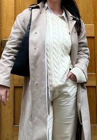 Bež trench coat kerge tekstuuriga sviitriga all, kreemivärvi püksid ja must käekott. Stiliseeritud puidust tausta ees.