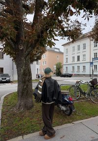 Giacca di pelle nera, maglione di lana beige, pantaloni larghi marroni, berretto verde e una sciarpa a righe, in piedi accanto a un albero con foglie marroni.