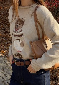 Béžový pletený sveter s motívom medveďa, hnedými detailmi a rebrovaným výstrihom. Kombinovaný s tmavými denimovými džínsami a hnedou kabelkou.