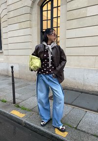 Veste surdimensionnée en cuir marron, haut à pois marron, pull gris noué au cou, jean large bleu clair et sac à main vert citron. Chaussures blanches et marron.