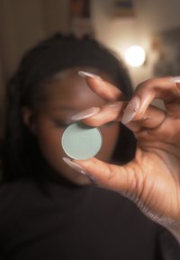 Une palette cosmétique ronde de couleur vert menthe tenue entre des doigts, présentant une surface texturée. L'arrière-plan est flou avec un éclairage doux.