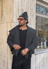 Homme tatoué portant un bonnet noir et un manteau, des lunettes de soleil ambrées et des bagues, debout près d'un mur en pierre beige et d'une vitrine de salon avec des produits capillaires.