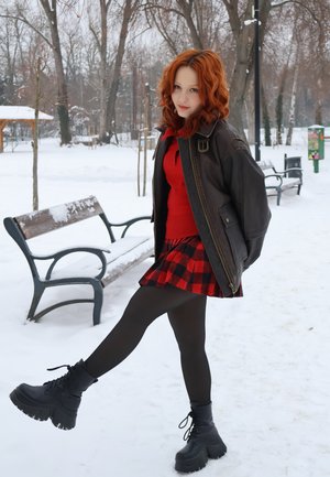 Joven con cabello rojo, chaqueta de cuero, suéter rojo y falda de cuadros levantando la pierna en un parque nevado con bancos y árboles sin hojas.