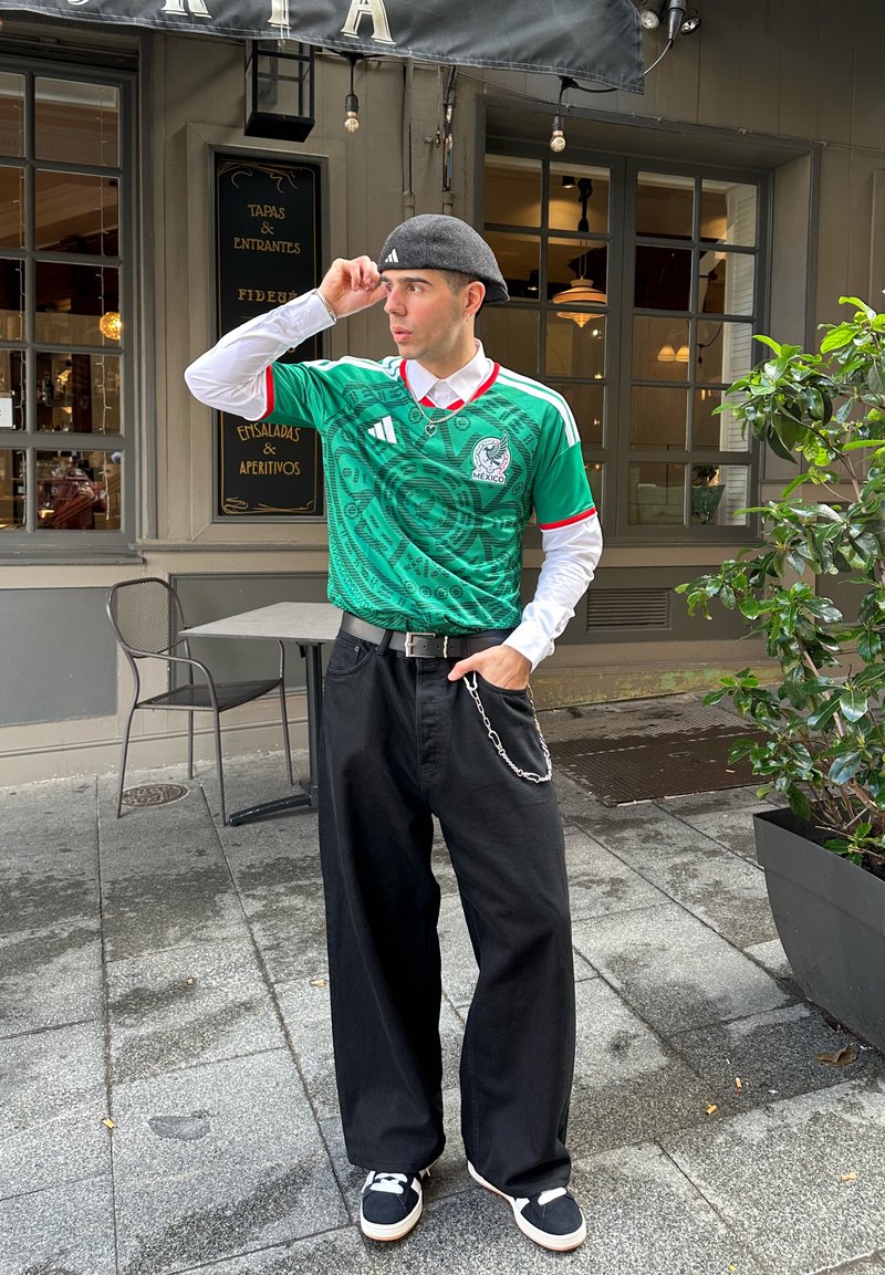 Jeune homme portant un maillot de football vert du Mexique, une chemise blanche à manches longues, un pantalon large noir, une casquette grise et des baskets, debout devant un café.