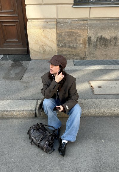 Joven mujer con sombrero y chaqueta marrones sentada en la acera de la calle ajustando los auriculares, sosteniendo el teléfono, con una bolsa negra a su lado.