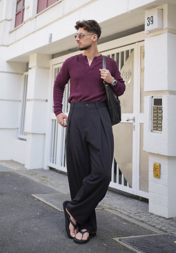 Homme portant des lunettes de soleil, une chemise à manches longues bordeaux, un pantalon large noir et des tongs, debout sur un trottoir en ville près d'un bâtiment blanc.