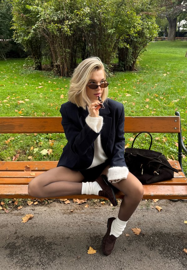 Blazer de rayas marineras con puños de piel blanca, suéter blanco, pantalones cortos negros, medias transparentes y mocasines marrones; accessorios incluyendo gafas de sol y un bolso negro.