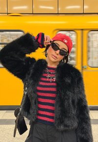 Veste en fausse fourrure noire, haut en maille rayé rouge et noir, casquette rouge, lunettes de soleil noires, collier argenté avec breloques, sur un fond jaune.