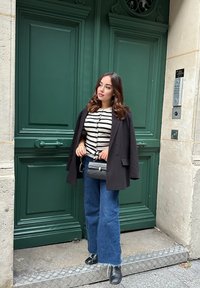 Schwarzer Blazer über einem gestreiften Pullover, kombiniert mit weit geschnittenen Denim-Jeans. Accessoires sind eine schwarze Handtasche und Sonnenbrille. Grüne Türen im Hintergrund.