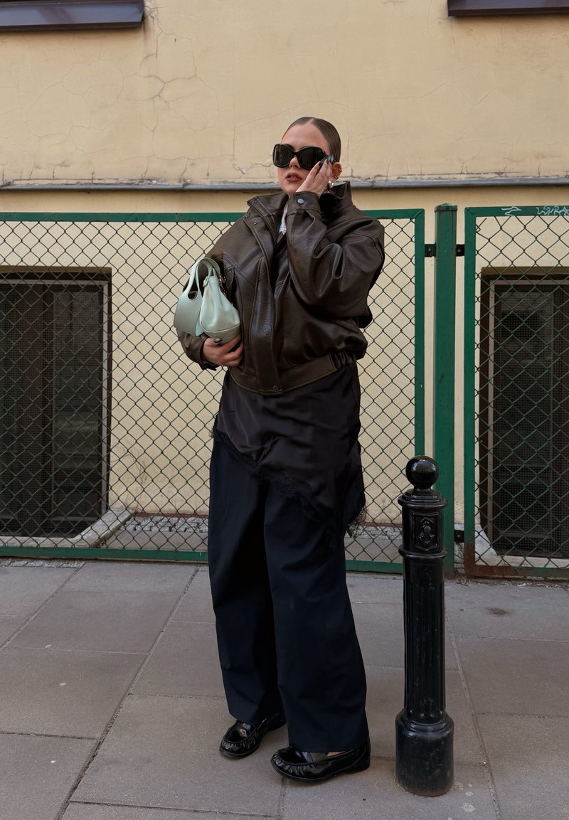 Personne portant une veste en cuir sombre surdimensionnée et un pantalon large tient un sac à main vert clair, porte de grandes lunettes de soleil, debout sur le trottoir de la ville près d'une clôture.