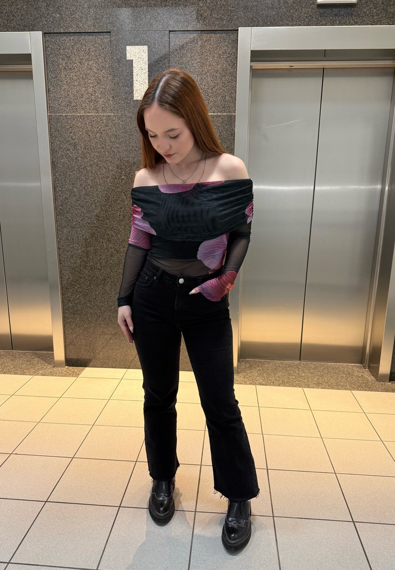 Jeune femme debout entre deux portes d'ascenseur, vêtue d'un haut fleuri noir sans épaules, un jean noir et des chaussures noires, regardant vers le bas.