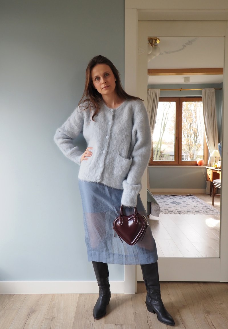 Eine Frau trägt eine flauschige, hellblaue, geknöpfte Strickjacke und einen durchsichtigen blauen Rock, hält eine herzförmige burgunderfarbene Handtasche und steht drinnen in der Nähe eines Spiegels.
