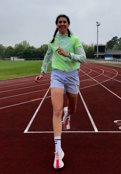 Corredora femenina con chaqueta verde y pantalones cortos azul claros sonriendo mientras corre en una pista exterior roja, con un campo verde y árboles de fondo.