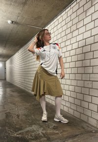 Weisssporttrikot mit schwarzen und roten Akzenten, khakifarbener asymmetrischer Rock, weisse Kniestrümpfe und hellgraue Sneakers. Innenraum mit Ziegelwänden.