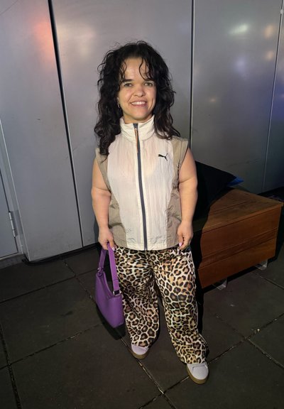 Persona vestida con un chaleco sin mangas color beige con cremallera y un logo negro, combinado con pantalones de estampado de leopardo y un bolso morado, de pie sobre la acera.