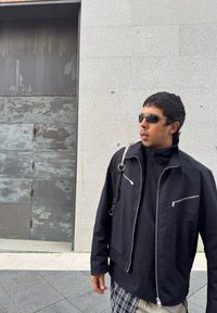 Μαύρο ελαφρύ μπουφάν με φερμουάρ, συνδυασμένο με μαύρη μπλούζα και εντύπωση παντελόνι. Τα αξεσουάρ περιλαμβάνουν γυαλιά ηλίου και τσάντα ώμου.