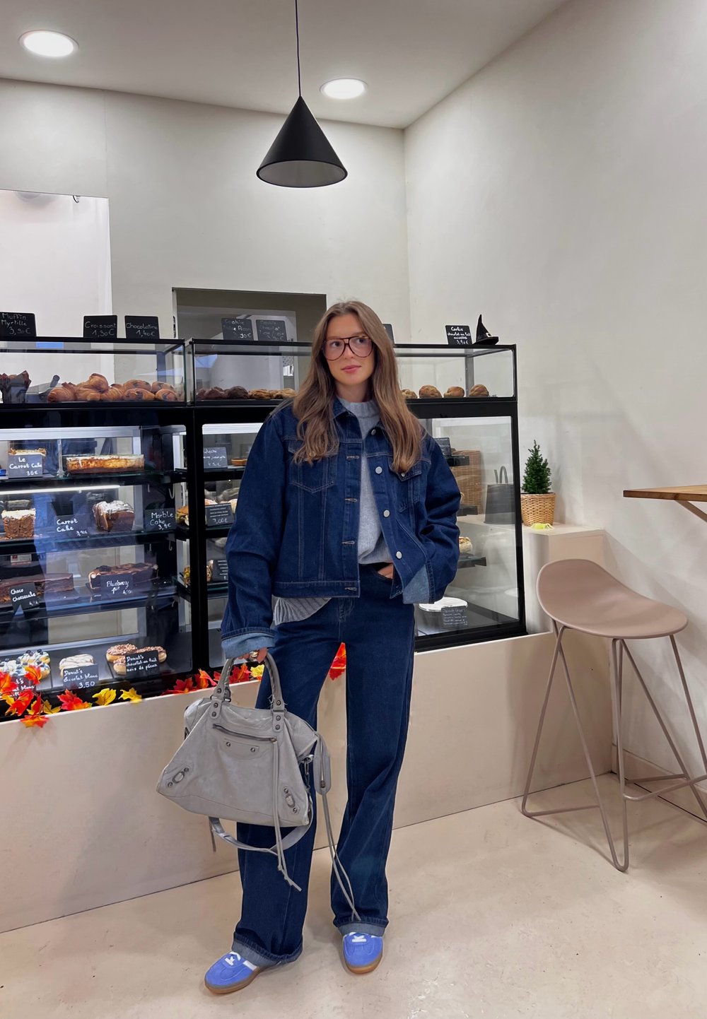 Mėlyna džinsinė striukė ir derančios kelnės, pilkas megztinis, pilna rankinė, mėlyni batai. Interjere yra vitrina su kepiniais ir neutralūs tonai.