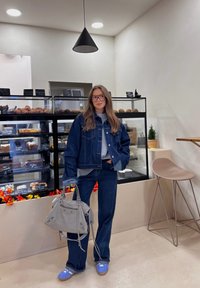 Mėlyna džinsinė striukė ir derančios kelnės, pilkas megztinis, pilna rankinė, mėlyni batai. Interjere yra vitrina su kepiniais ir neutralūs tonai.