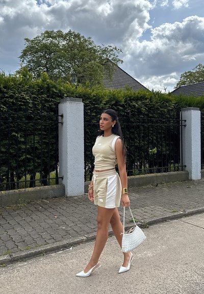 Top corto de color beige claro y pantalones cortos a juego con paneles laterales blancos. Zapatillas de tacón blanco y un bolso de mano tejido blanco. Entorno al aire libre.
