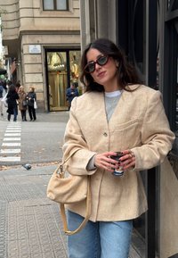 Jeune femme en blazer beige et lunettes de soleil tenant une tasse de café, debout sur un trottoir en ville près de l'entrée d'un bâtiment.