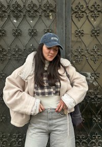 Light beige oversized fleece jacket with a white lining, layered over a checked shirt. Grey high-waisted jeans and a navy cap.