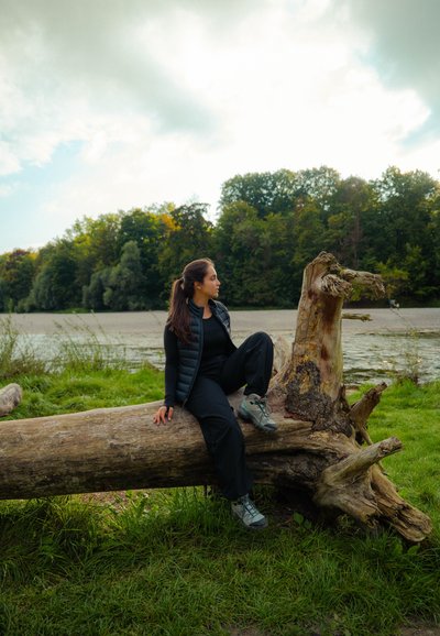 Una mujer vestida con ropa negra de outdoor se sienta en un tronco de árbol caído cerca de una ribera de hierba, con un bosque y un cielo nublado de fondo.