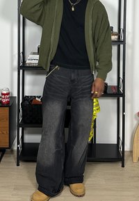 Olivengrüner, gerippter Zip-Up-Hoodie, schwarzes T-Shirt, ausgebleichte schwarze weit geschnittene Jeans, braune Stiefel und mehrlagige goldene Halsketten mit Ketten und Anhängern.