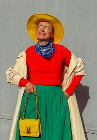 Rotes Langarmoberteil, grüner plissierter Rock, gelbe Handtasche mit goldenen Beschlägen, gelber Hut und blaues gemustertes Tuch. Helle Jacke drapiert.