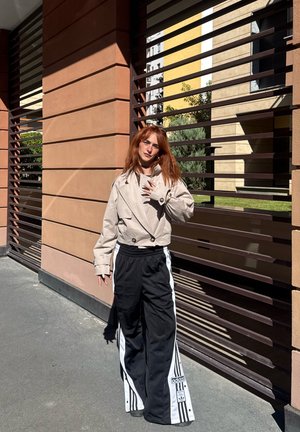 Veste croisée beige coupée avec de grands boutons, associée à un pantalon de sport noir avec des rayures blanches et un logo, sur fond de lattes.