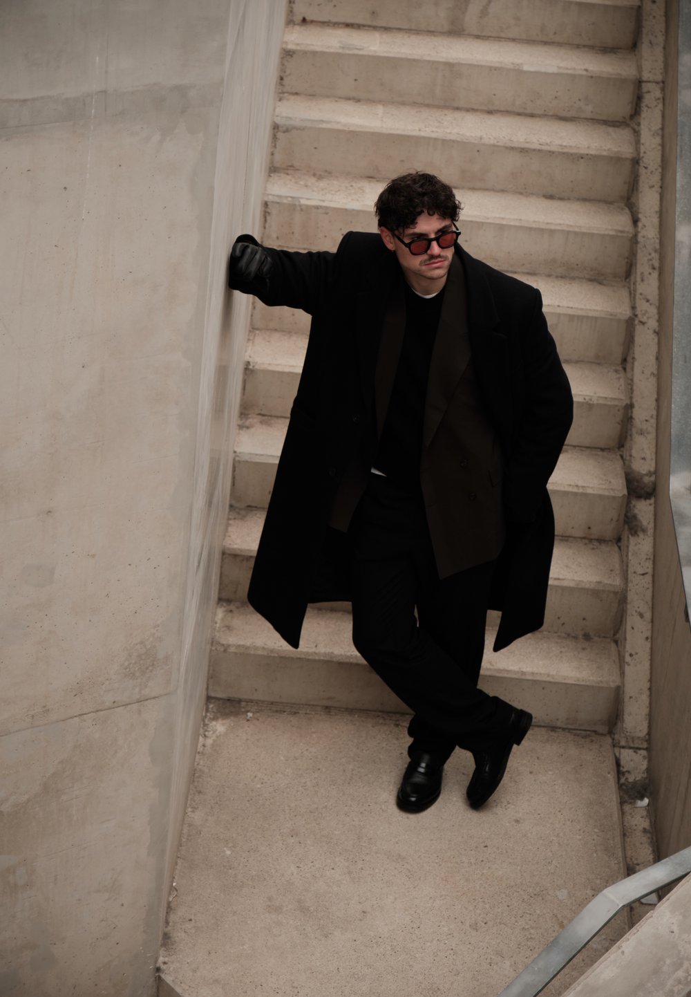 Homme portant un manteau noir, des lunettes de soleil et des gants, appuyé contre un mur en béton au bas d'un escalier, les jambes croisées.