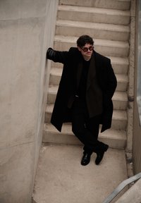 Homme portant un manteau noir, des lunettes de soleil et des gants, appuyé contre un mur en béton au bas d'un escalier, les jambes croisées.