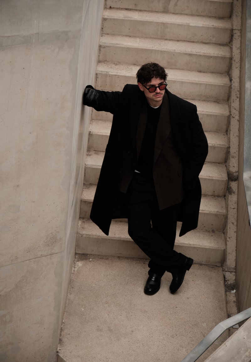 Uomo con cappotto nero, occhiali da sole e guanti, appoggiato a un muro di cemento in fondo a una scala con le gambe incrociate.