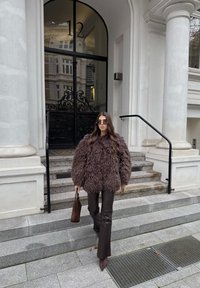 Palton maro pufos, pantaloni mulați din piele maro și geantă maro. Stând pe trepte de piatră în fața unei uși negre din fier forjat.