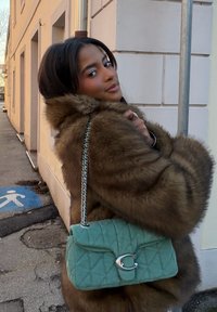 Manteau en fourrure brune avec un grand col ; portant un sac à main vert matelassé avec une fermeture argentée. Cadre urbain avec trottoir et bâtiment en arrière-plan.