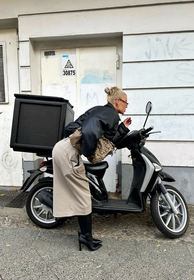 Patinete negro con una caja de entrega rectangular, pavimento gris, y una mujer con chaqueta negra, falda beige y botas de tacón negro.