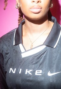 Chemise de sport noire avec col, arborant des logos et des accents Nike blancs. Tissu brillant avec un aspect texturé. Collier en chaîne doré visible.