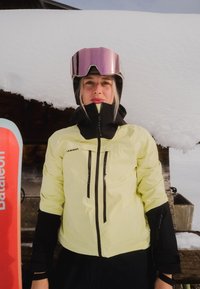 Femme en veste de ski jaune et lunettes de protection roses réfléchissantes se tient devant une cabine enneigée, avec un ski rouge visible à gauche.