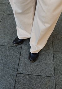 Schwarze Lederschuhe mit runder Zehenform, die gelbe Nähte entlang der Sohle aufweisen, getragen mit lässigen beige Hosen auf einem steinernen Untergrund.