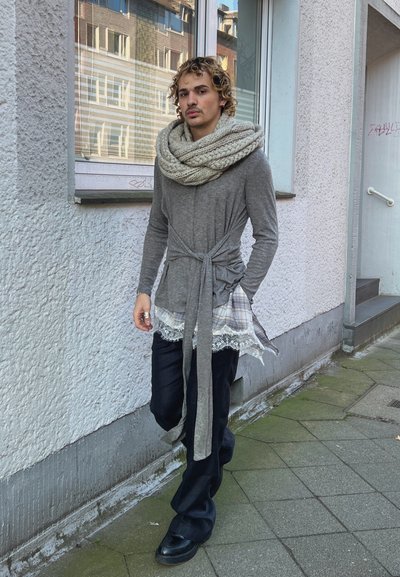 Hombre con cabello rizado que lleva una bufanda gruesa de punto, camisa de manga larga gris con cinturón sobre una blusa de cuadros con encaje, pantalones negros, de pie en una acera de la ciudad.