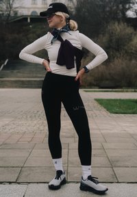 Zwarte legging met klein logo, crème lange mouwen top, paarse trui vastgebonden in de taille, en grijze schoenen met zwarte accenten.